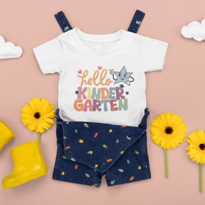 May include: White baby onesie with a rainbow colored graphic that says "hello kindergarten" with a cartoon star. The onesie is paired with blue denim overalls with a bug pattern.