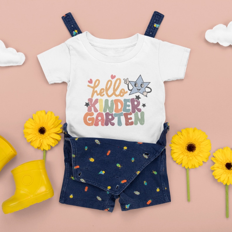 May include: White baby onesie with a rainbow colored graphic that says "hello kindergarten" with a cartoon star. The onesie is paired with blue denim overalls with a bug pattern.