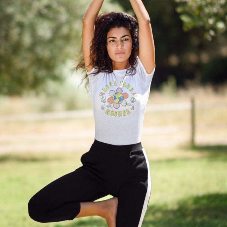 May include: A woman wearing a white t-shirt with a flower graphic and the text "Love Your Mother" in a retro font. She is wearing black pants with a white stripe down the side and is standing in a yoga pose.