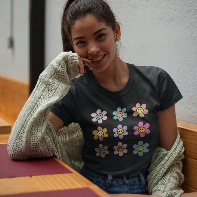 May include: A black t-shirt featuring a colourful pattern of smiley face flowers. The flowers are in shades of pink, blue, yellow, and green.