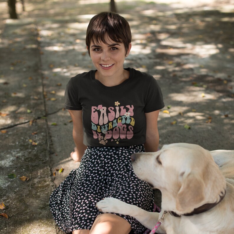 May include: A woman wearing a black t-shirt with the text "Easily Distracted by Dogs" in a retro font with floral accents. She is sitting on the ground with a white dog.