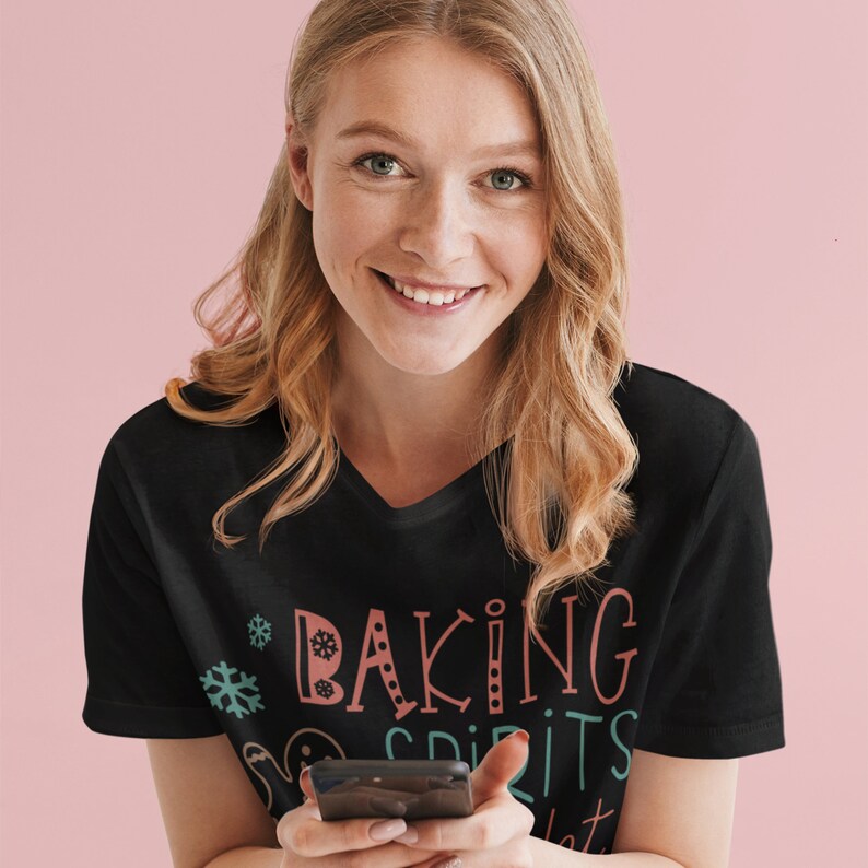 May include: A woman wearing a black t-shirt with the text "Baking Spirits Bright" in colorful lettering. She is holding a black smartphone in her hands.