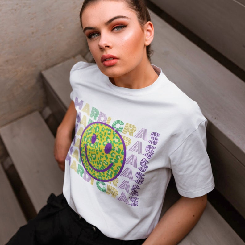 May include: White t-shirt with a repeating "MARDI GRAS" design. The design includes a yellow smiley face with a leopard print pattern and purple eyes. The text is in green, purple, and yellow.
