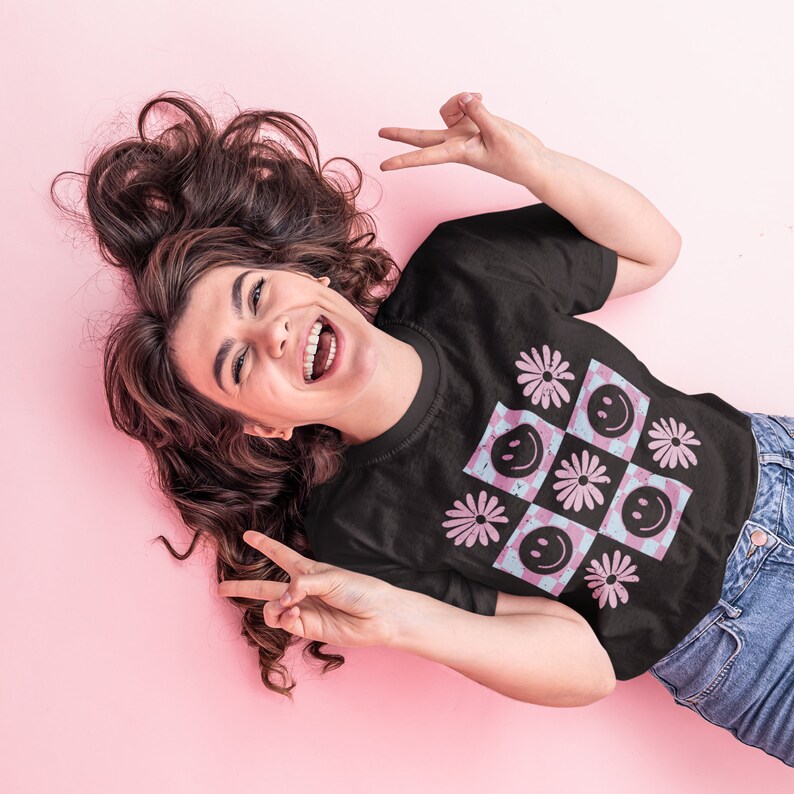 Pode incluir: Uma camiseta preta com um padr&atilde;o xadrez rosa e azul com flores rosa e rostos sorridentes.