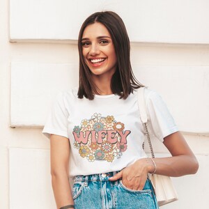 May include: White t-shirt with a pink, blue, and yellow floral design and the word "WIFEY" in pink letters.