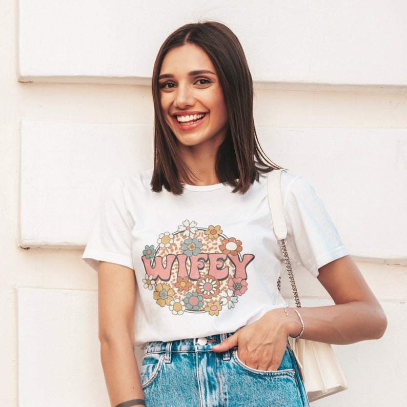 May include: White t-shirt with a pink, blue, and yellow floral design and the word "WIFEY" in pink letters.