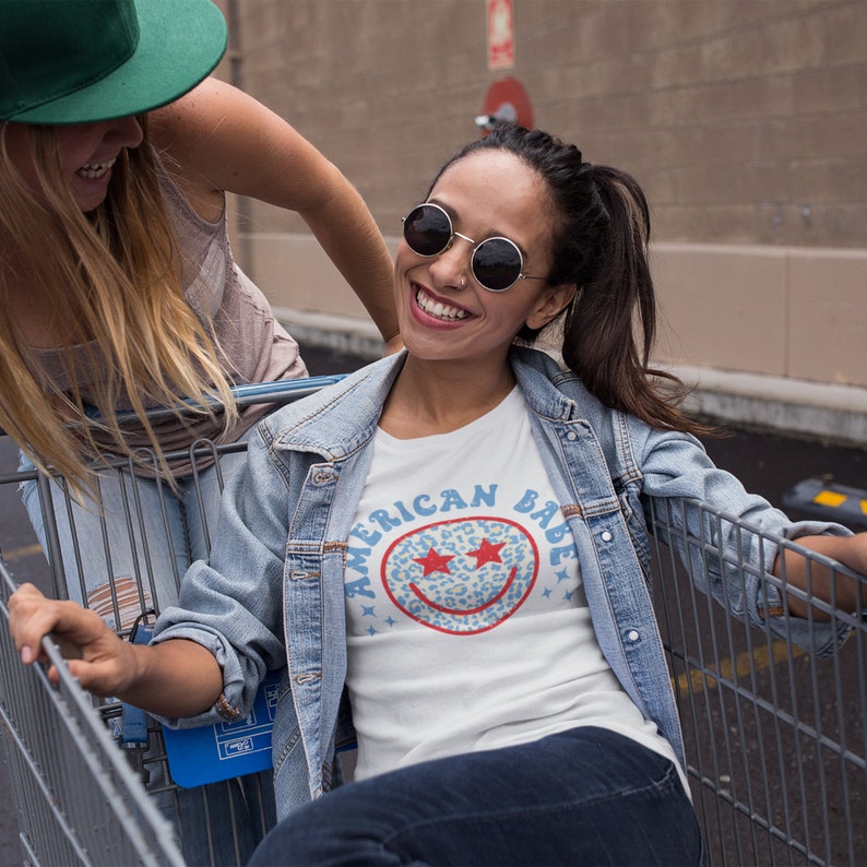 May include: Two friends are riding in a shopping cart. The friend in the front is wearing a white t-shirt with a blue and red smiley face graphic that says "American Babe". She is also wearing a denim jacket and sunglasses.