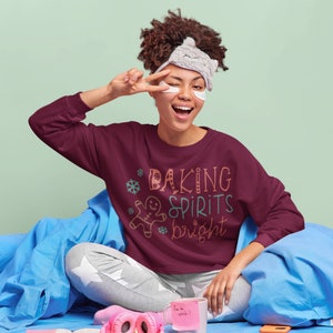 May include: A maroon sweatshirt with the text "Baking Spirits Bright" in colorful lettering and a gingerbread man graphic. The sweatshirt is worn by a person with a sleep mask and eye patches.
