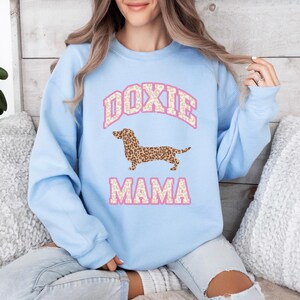 May include: Light blue sweatshirt with a pink "Doxie Mama" graphic featuring a dachshund with a leopard print pattern.