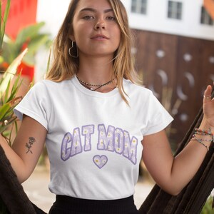 May include: White t-shirt with a purple "Cat Mom" graphic with a daisy pattern. The graphic has a small heart with a daisy pattern below it.