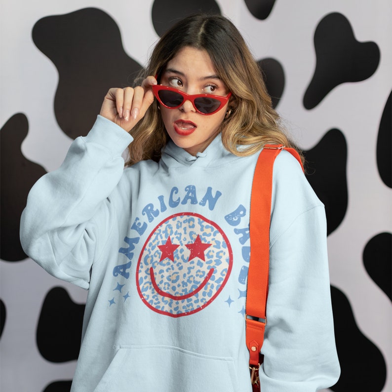 May include: A light blue hoodie with a distressed graphic of a smiley face with stars for eyes and a leopard print design. The text "AMERICAN BAD" is written around the smiley face.