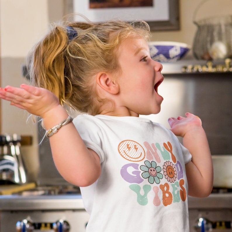 May include: A young girl wearing a white t-shirt with a colorful graphic that says "Only Good Vibes". The graphic features a smiley face, a flower, and a sun.