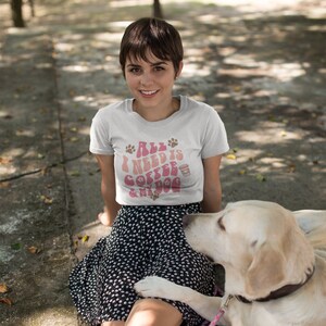 Pu&ograve; includere: Una maglietta bianca con la scritta "All I Need Is Coffee & My Dog" in rosa e marrone. La maglietta presenta impronte di zampe e una grafica a tazza di caff&egrave;. La persona &egrave; seduta fuori con un cane.