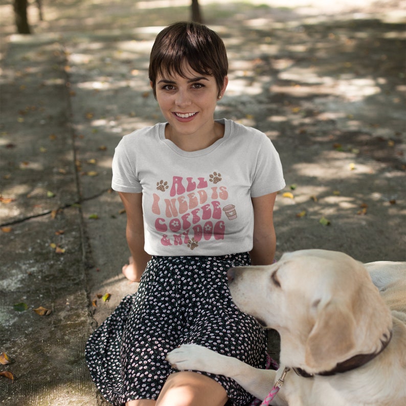 Pu&ograve; includere: Una maglietta bianca con la scritta "All I Need Is Coffee & My Dog" in rosa e marrone. La maglietta presenta impronte di zampe e una grafica a tazza di caff&egrave;. La persona &egrave; seduta fuori con un cane.