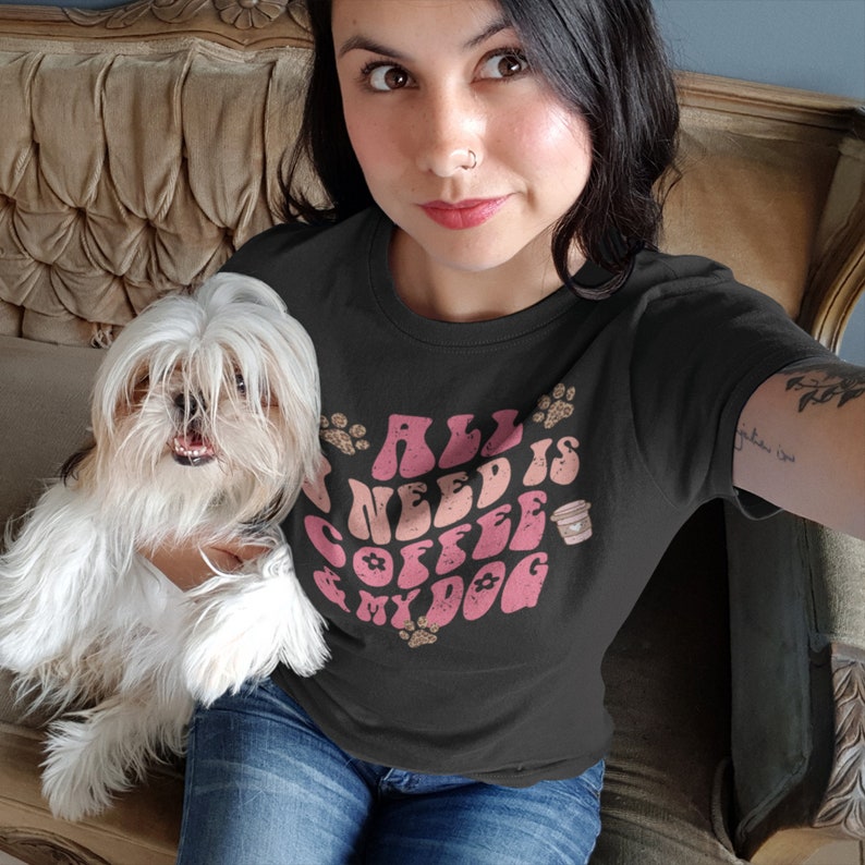 Pu&ograve; includere: Una maglietta nera con la scritta "ALL I NEED IS COFFEE & MY DOG" in lettere rosa e pesca. La maglietta presenta impronte di zampe e una grafica di tazza di caff&egrave;. La persona indossa jeans blu e tiene in braccio un cagnolino bianco.
