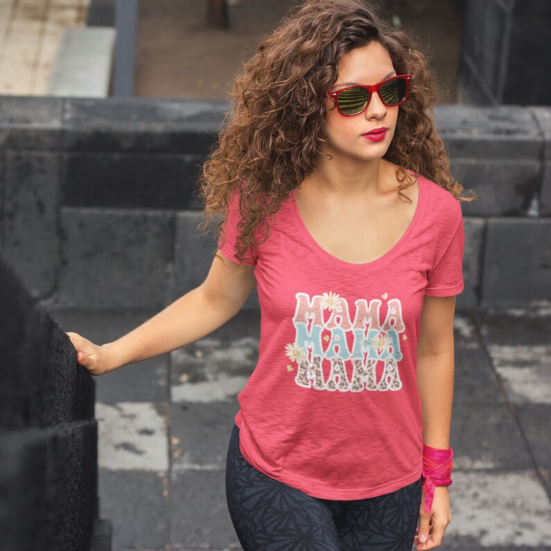 Puede incluir: Una camiseta rosa de cuello redondo con el texto "Mama Mama Mama" en una fuente superpuesta y colorida con acentos florales y estampados de animales.