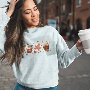 May include: A light blue sweatshirt featuring a graphic of autumnal drinks and leaves. The design includes a pumpkin spice latte, a hot chocolate, and a coffee drink. The sweatshirt is being worn by a person holding a white takeaway cup.