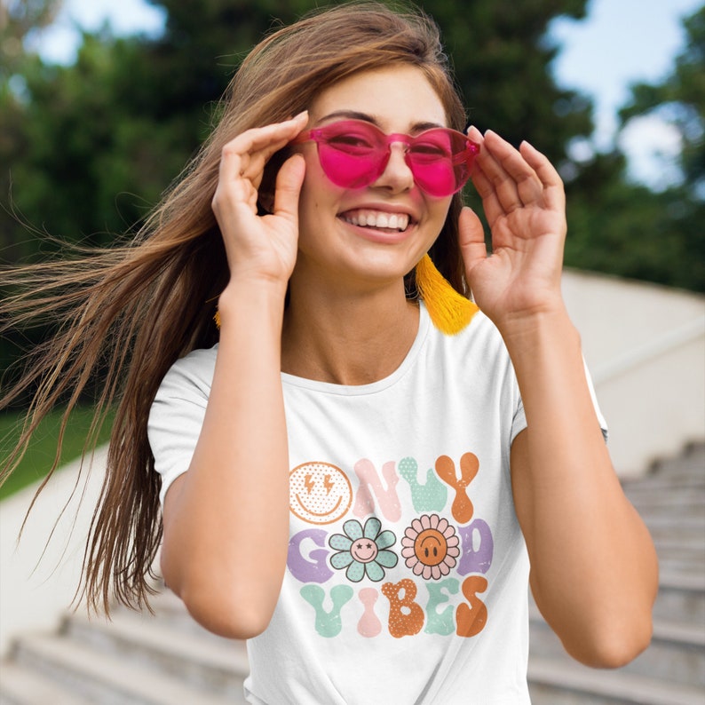 May include: White t-shirt with a colorful graphic that says "Only Good Vibes" with a smiley face, flower, and sun design.