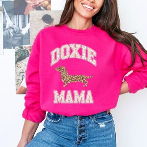 May include: Pink sweatshirt with a white and brown dachshund graphic and the text "Doxie Mama" in a varsity style font.