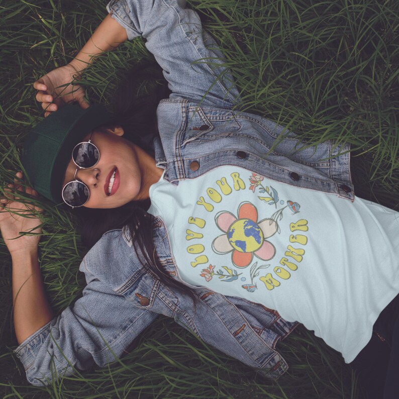 May include: A light blue t-shirt with a flower graphic and the text "Love Your Mother" in a retro style. The flower has a pink, orange, and yellow center with a green and blue globe in the middle. The shirt is worn by a person lying in a field of green grass.