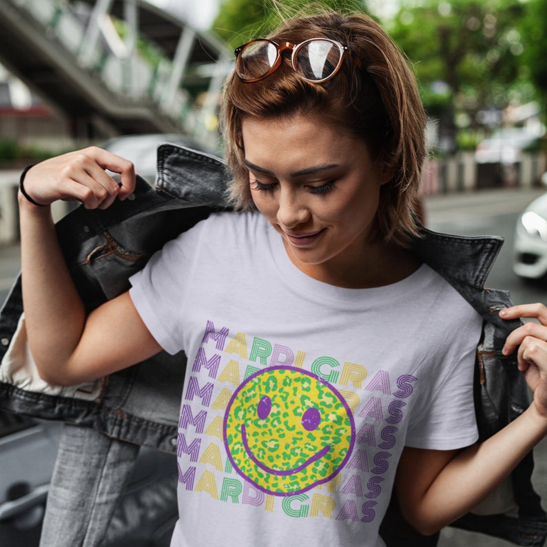 May include: White t-shirt featuring a smiling face with a leopard print design in yellow and green. The word "MARDI GRAS" is printed in a repeating pattern in purple and green. The model is wearing a denim jacket.