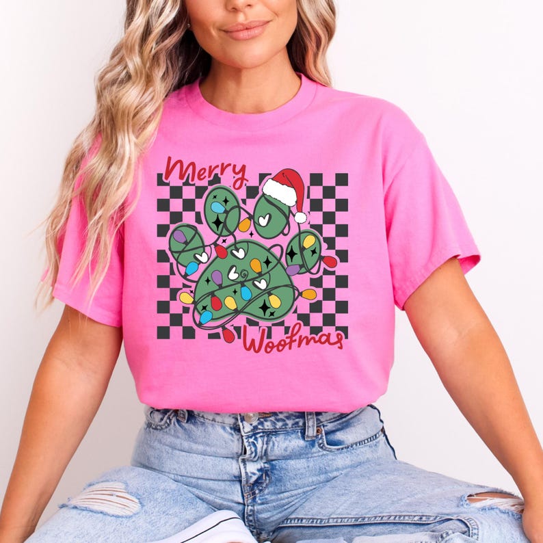 Puede incluir: Camiseta rosa con un dise&ntilde;o de huella de pata verde con luces de Navidad y un gorro de Pap&aacute; Noel. El texto "Merry Woofmas" est&aacute; impreso en la camiseta.
