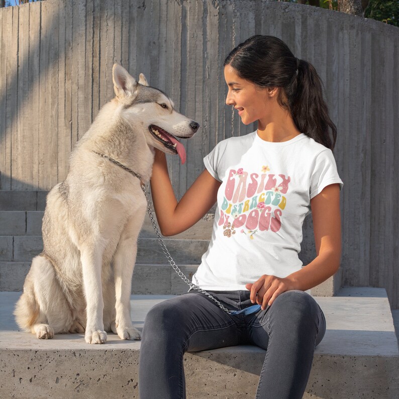 May include: A woman wearing a white t-shirt with the text "Easily Distracted by Dogs" in a retro font with floral accents. She is petting a white and grey husky dog.