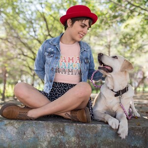 Puede incluir: Una mujer con una camiseta rosa con el texto "Mama Mama" en colores arco&iacute;ris y un sombrero rojo se sienta con las piernas cruzadas. Est&aacute; acariciando a un perro blanco con una correa rosa.