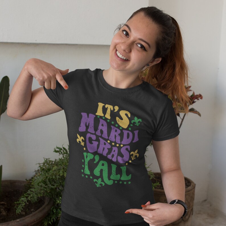 K&ouml;nnte beinhalten: Ein schwarzes T-Shirt mit einer lila, gr&uuml;nen und gelben Grafik, auf der "It's Mardi Gras Y'all" steht.