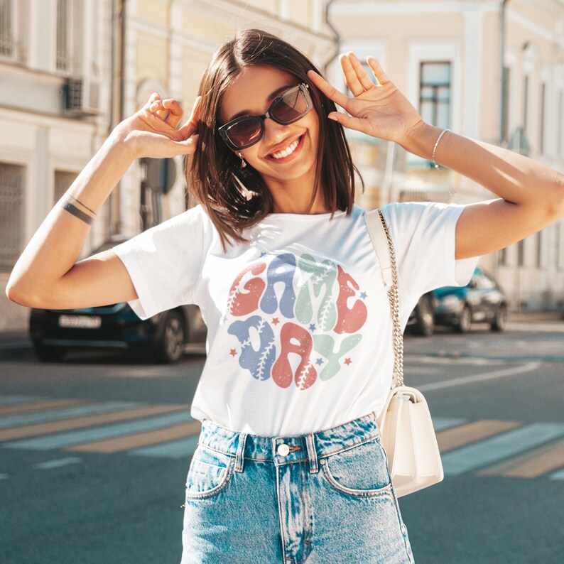 May include: A white t-shirt featuring a colourful graphic of a baseball with the text "GAME DAY" inside. The graphic is in a retro style with a distressed look.