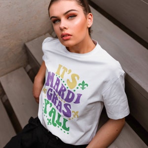 K&ouml;nnte beinhalten: Wei&szlig;es T-Shirt mit dem Text "It's Mardi Gras Y'all" in lila, gelber und gr&uuml;ner Schrift mit gr&uuml;nen Fleur-de-lis-Akzenten.
