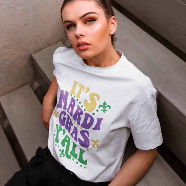 K&ouml;nnte beinhalten: Wei&szlig;es T-Shirt mit dem Text "It's Mardi Gras Y'all" in lila, gelber und gr&uuml;ner Schrift mit gr&uuml;nen Fleur-de-lis-Akzenten.