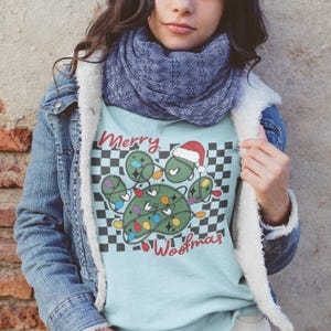 Puede incluir: Una camiseta azul claro con un dise&ntilde;o de huella de pata verde. La huella de pata est&aacute; decorada con luces de Navidad y un gorro de Pap&aacute; Noel. El texto "Merry Woofmas" est&aacute; impreso debajo de la huella de pata.