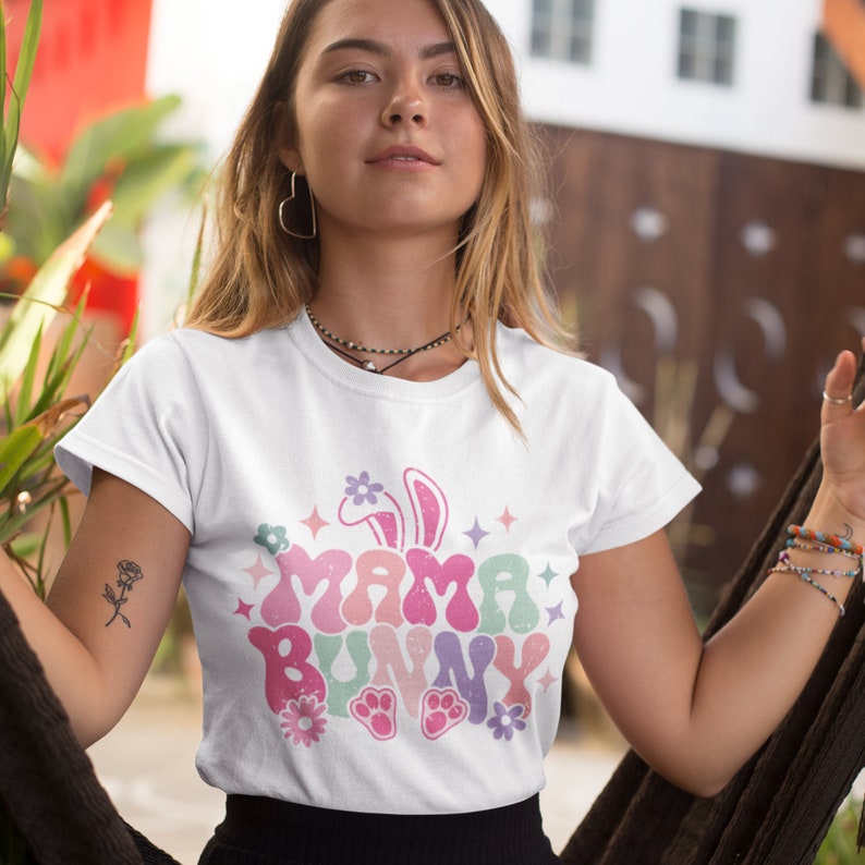 May include: White t-shirt featuring a colourful graphic design that reads "Mama Bunny" in a retro style. The design includes a bunny rabbit with ears, flowers, and paw prints.