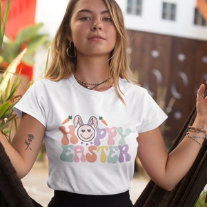 May include: White t-shirt with a colorful graphic that says "Hoppy Easter" with a cartoon bunny wearing a pink and white checkered bow. The bunny has a smiling face with two hearts for eyes.