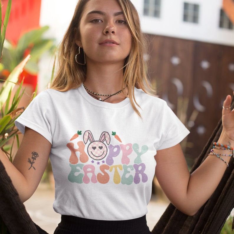 May include: White t-shirt with a colorful graphic that says "Hoppy Easter" with a cartoon bunny wearing a pink and white checkered bow. The bunny has a smiling face with two hearts for eyes.