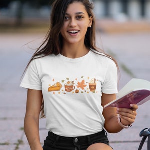 May include: White t-shirt featuring a graphic of a pumpkin spice latte, a slice of pumpkin pie, and an autumn leaf. The text on the latte reads "Pumpkin Spice Latte".