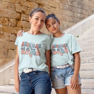 May include: Two people wearing light blue t-shirts with the text "American Mama" and "American Babe" in red, white, and blue. The text is stylized with a retro font and features a small American flag design.