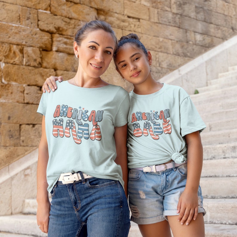 May include: Two people wearing light blue t-shirts with the text "American Mama" and "American Babe" in red, white, and blue. The text is stylized with a retro font and features a small American flag design.