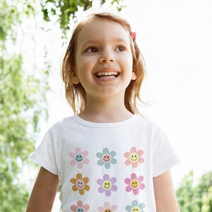 May include: A young girl with long brown hair smiles while wearing a white t-shirt with a pattern of colourful smiley face flowers.