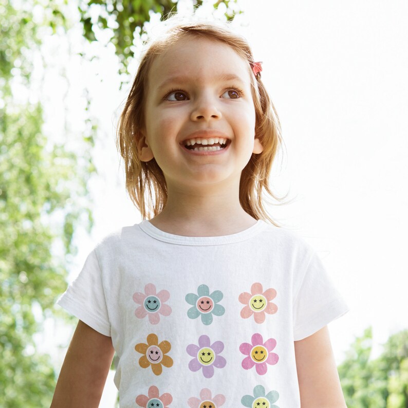 May include: A young girl with long brown hair smiles while wearing a white t-shirt with a pattern of colourful smiley face flowers.