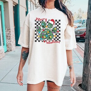 Puede incluir: Una camiseta blanca con un dise&ntilde;o de huella de pata con tem&aacute;tica navide&ntilde;a. La huella de pata es verde y tiene un gorro de Pap&aacute; Noel rojo en la parte superior. La huella de pata est&aacute; rodeada por un patr&oacute;n de cuadros negros y blancos. El texto "Merry Woofmas" est&aacute; escrito debajo de la huella de pata.