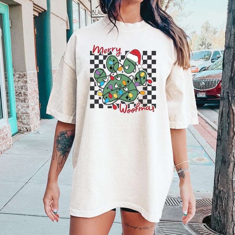 Puede incluir: Una camiseta blanca con un dise&ntilde;o de huella de pata con tem&aacute;tica navide&ntilde;a. La huella de pata es verde y tiene un gorro de Pap&aacute; Noel rojo en la parte superior. La huella de pata est&aacute; rodeada por un patr&oacute;n de cuadros negros y blancos. El texto "Merry Woofmas" est&aacute; escrito debajo de la huella de pata.