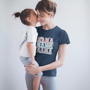 Puede incluir: Una mujer con una camiseta azul marino con el texto "MAMA" impreso en un dise&ntilde;o superpuesto y colorido sostiene a un ni&ntilde;o peque&ntilde;o. El ni&ntilde;o lleva una camiseta blanca y pantalones grises.
