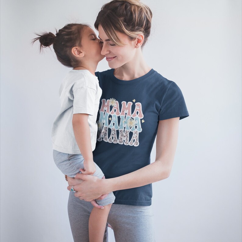Puede incluir: Una mujer con una camiseta azul marino con el texto "MAMA" impreso en un dise&ntilde;o superpuesto y colorido sostiene a un ni&ntilde;o peque&ntilde;o. El ni&ntilde;o lleva una camiseta blanca y pantalones grises.