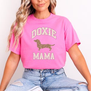 May include: Pink t-shirt with a white graphic of a dachshund dog with a leopard print pattern. The text "Doxie Mama" is printed below the dog.