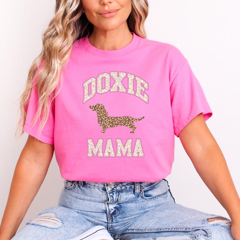 May include: Pink t-shirt with a white graphic of a dachshund dog with a leopard print pattern. The text "Doxie Mama" is printed below the dog.