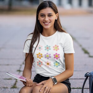 May include: A white t-shirt featuring a colourful pattern of flowers with smiley faces. The flowers are in shades of pink, yellow, blue, and green.