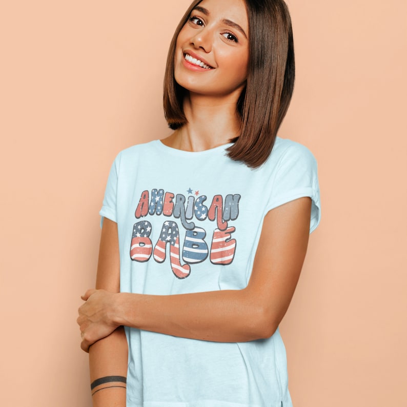May include: A light blue t-shirt with the text "AMERICAN BABE" in a retro font with red, white, and blue stars and stripes.