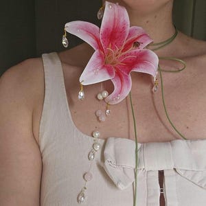 May include: A large pink and white lily flower brooch featuring dangling clear crystal teardrops and round white pearls. The floral accessory is attached to a thin green cord that wraps around the neck, accented with additional pearls and crystals.